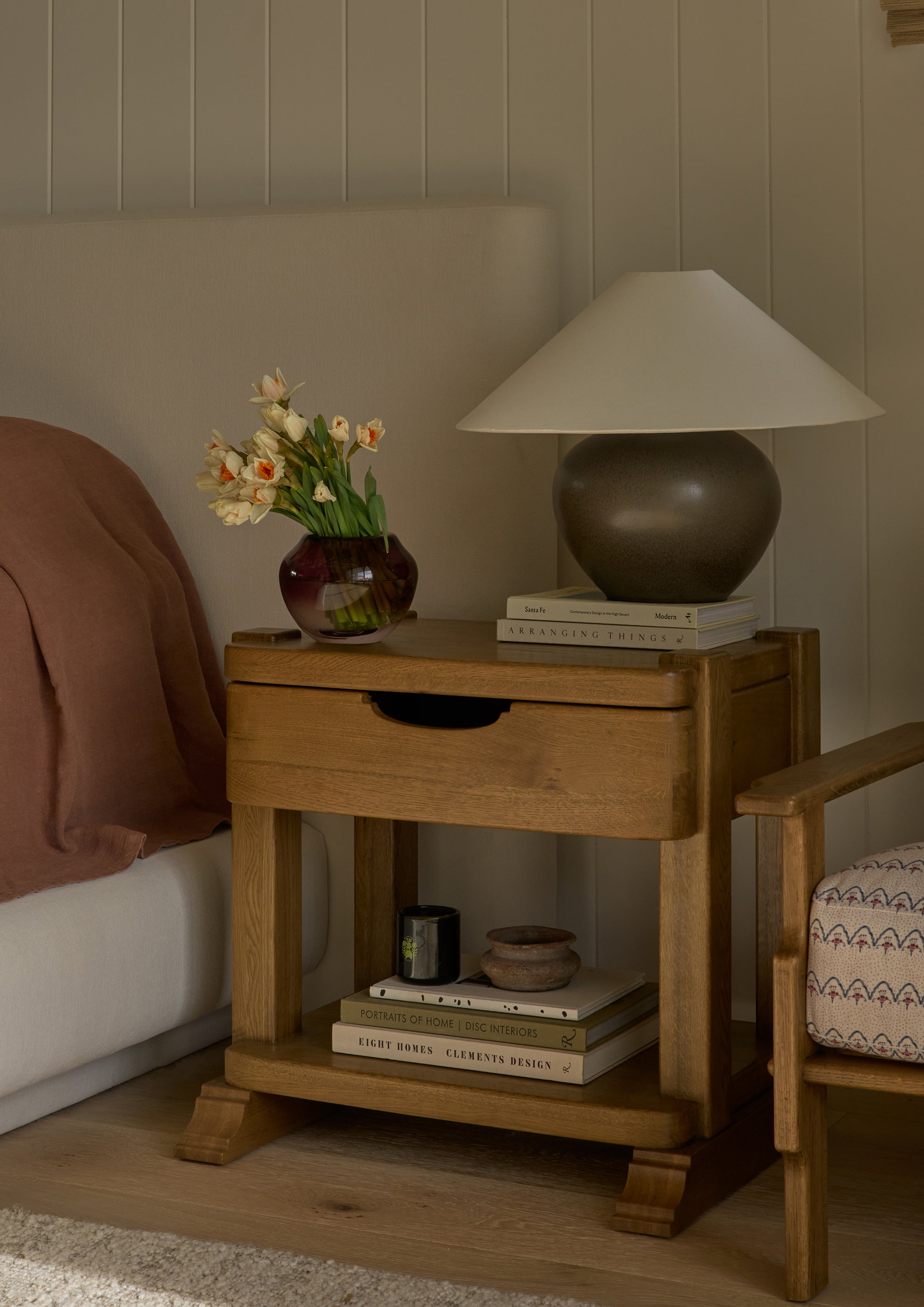 Honey oak nightstand styled beside an upholstered bed with a sculptural table lamp, vase of flowers, books, and decorative objects. The open shelf below provides additional storage while creating a warm and inviting bedside vignette.