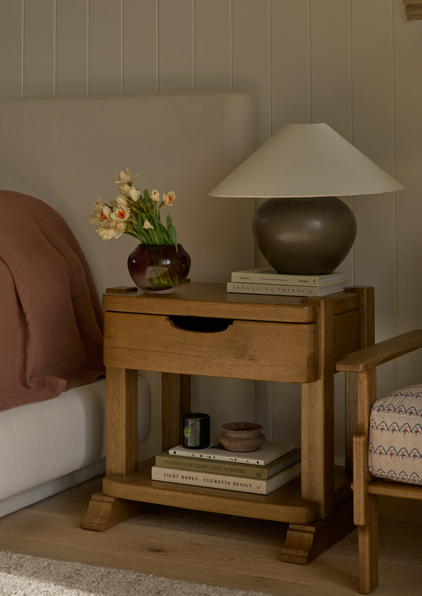 Honey oak nightstand styled beside an upholstered bed with a sculptural table lamp, vase of flowers, books, and decorative objects. The open shelf below provides additional storage while creating a warm and inviting bedside vignette.