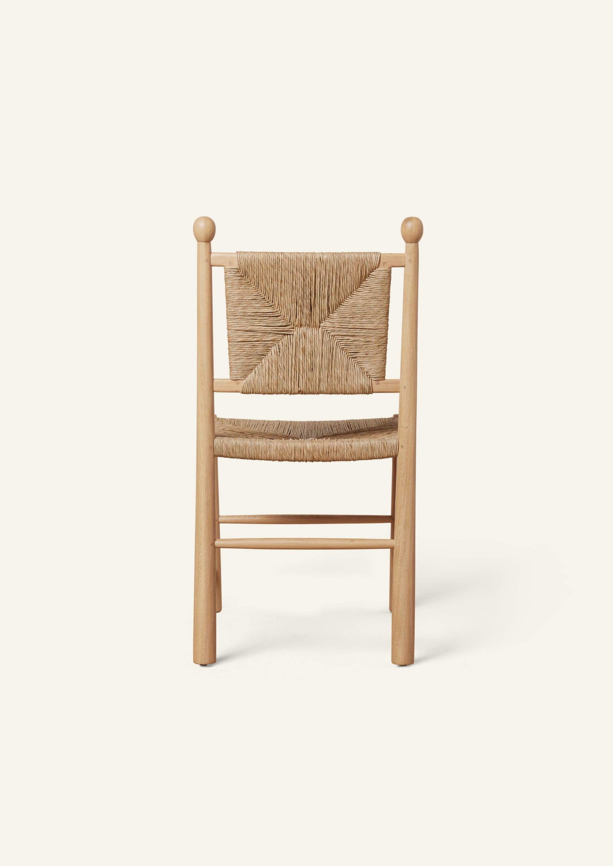 Rear view of a light oak dining chair with a handwoven rush seat and decorative woven backrest. Rounded back posts with turned finials frame the geometric rush pattern, while simple stretcher supports add stability. Photographed against a soft ivory background.