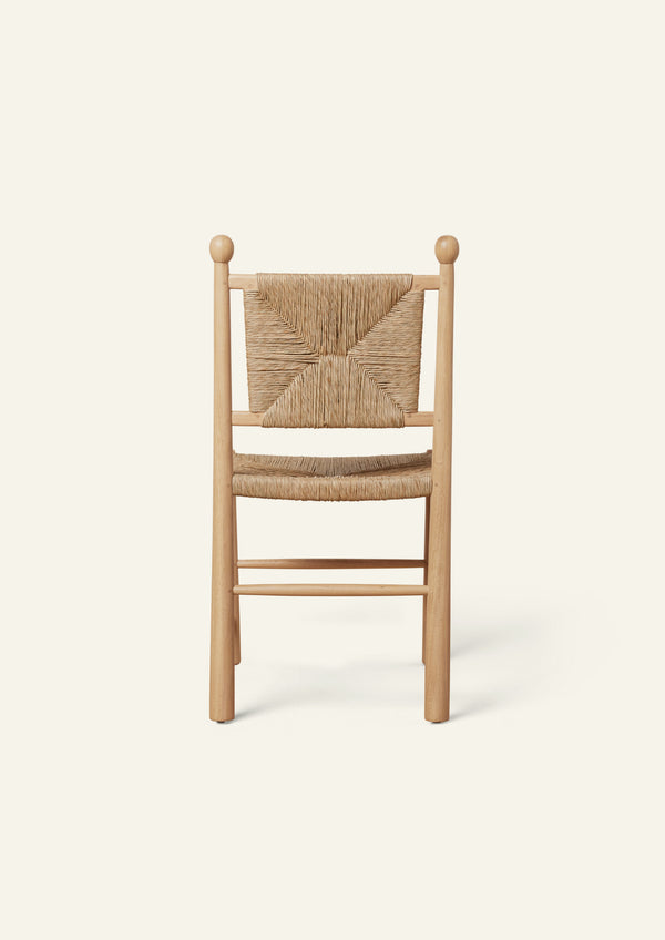Rear view of a light oak dining chair with a handwoven rush seat and decorative woven backrest. Rounded back posts with turned finials frame the geometric rush pattern, while simple stretcher supports add stability. Photographed against a soft ivory background.