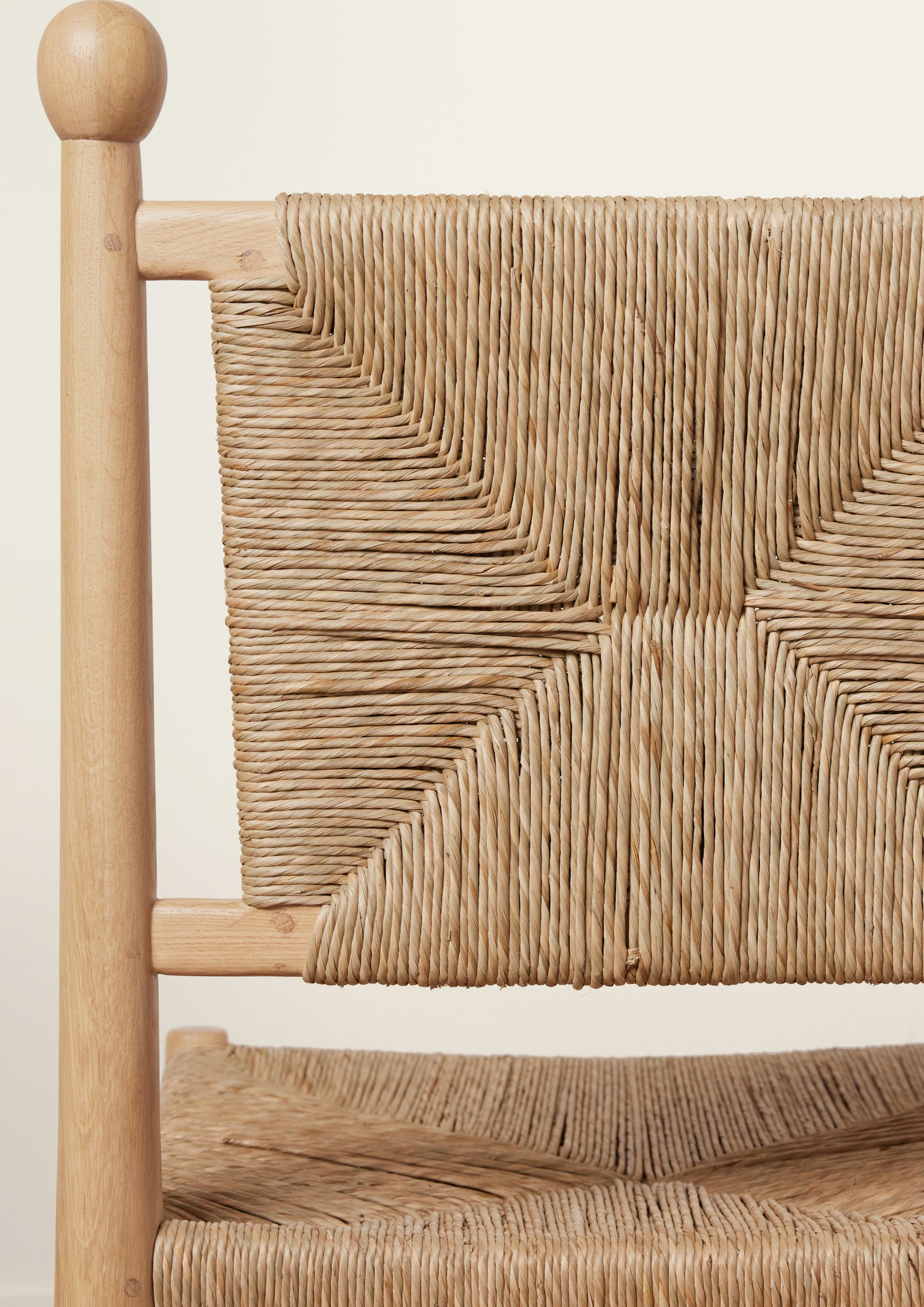 Close-up of a natural oak dining chair showcasing the handwoven rush backrest. The intricate geometric weave forms an hourglass pattern, framed by rounded wood posts topped with decorative finials. Part of the woven seat is visible below against a soft ivory background.