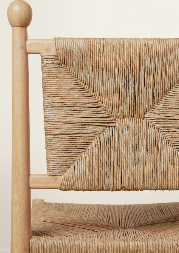 Close-up of a natural oak dining chair showcasing the handwoven rush backrest. The intricate geometric weave forms an hourglass pattern, framed by rounded wood posts topped with decorative finials. Part of the woven seat is visible below against a soft ivory background.