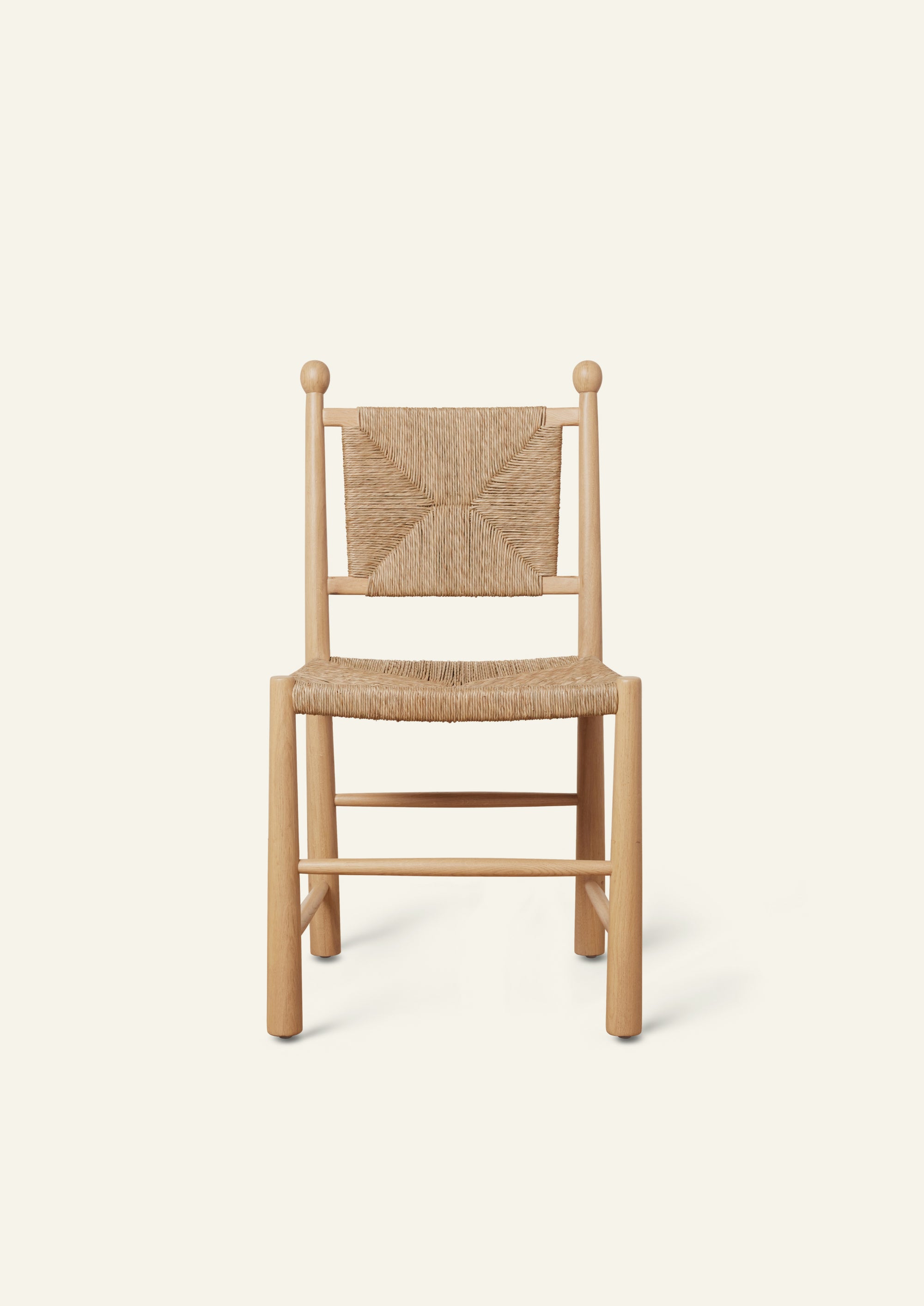 Front view of a light oak dining chair featuring a handwoven rush seat and geometric woven backrest. The chair has rounded legs, simple stretcher supports, and decorative finials atop the back posts, shown against a warm ivory backdrop.