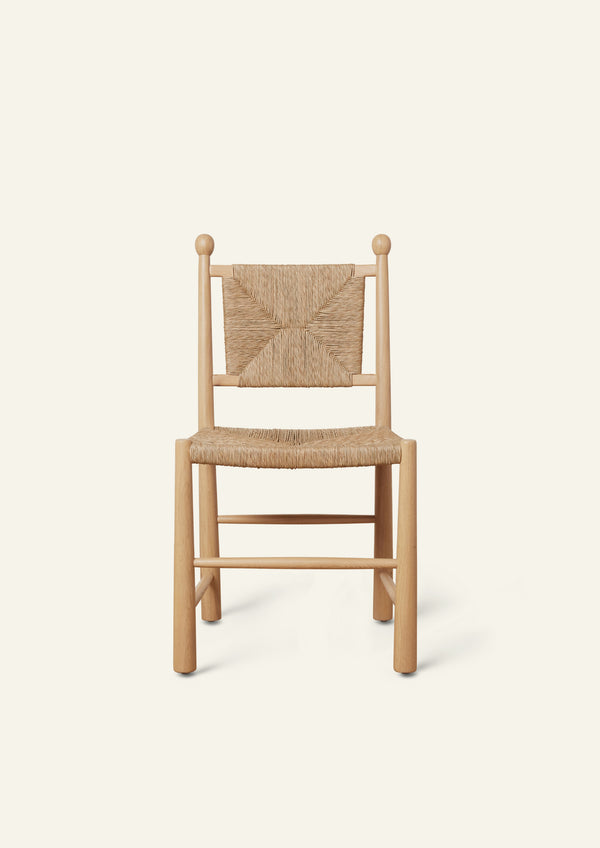 Front view of a light oak dining chair featuring a handwoven rush seat and geometric woven backrest. The chair has rounded legs, simple stretcher supports, and decorative finials atop the back posts, shown against a warm ivory backdrop.