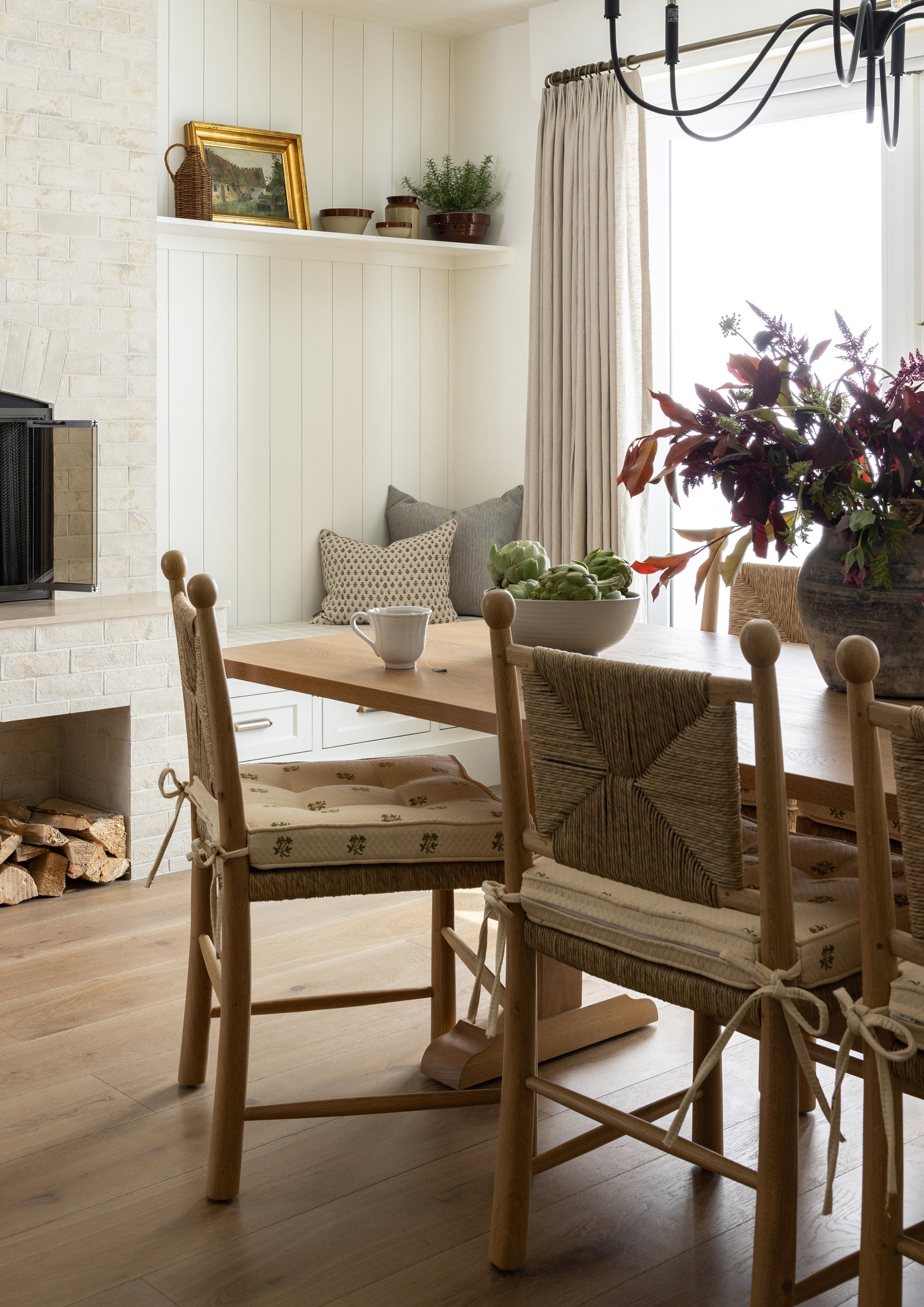 A set of natural oak dining chairs with handwoven rush seats and geometric woven backrests gathered around a wood dining table in a bright, traditional kitchen. Built-in cabinetry, soft window treatments, and a large arrangement of seasonal flowers create a warm, inviting atmosphere.