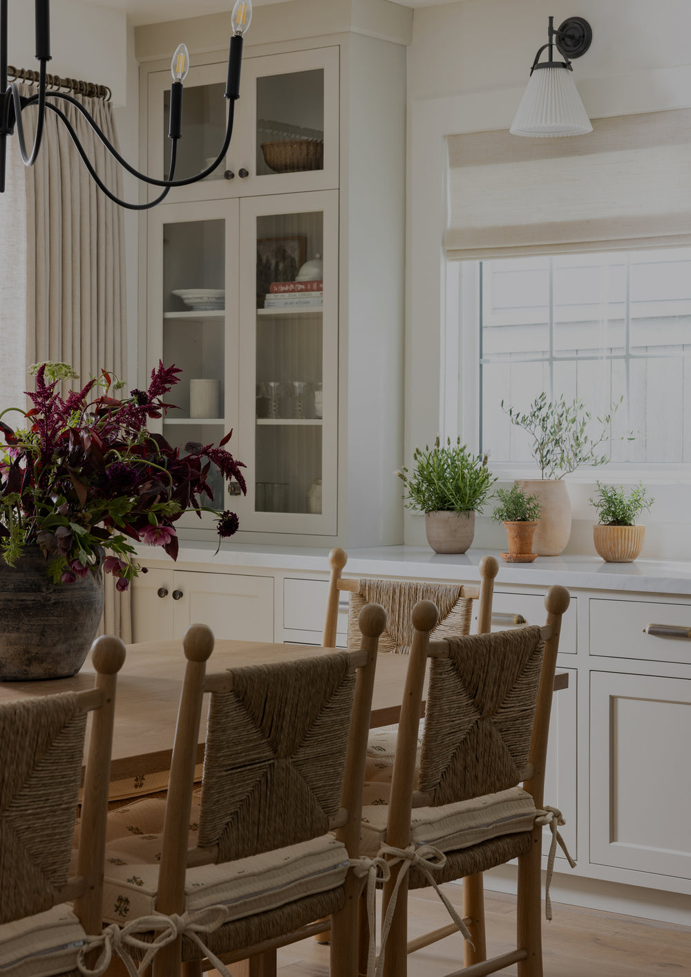 Natural oak dining chairs with woven rush seats and geometric woven backrests styled around a wood dining table in a light-filled dining nook. Floral seat cushions, a built-in bench, and a white brick fireplace contribute to the room’s relaxed, collected feel.