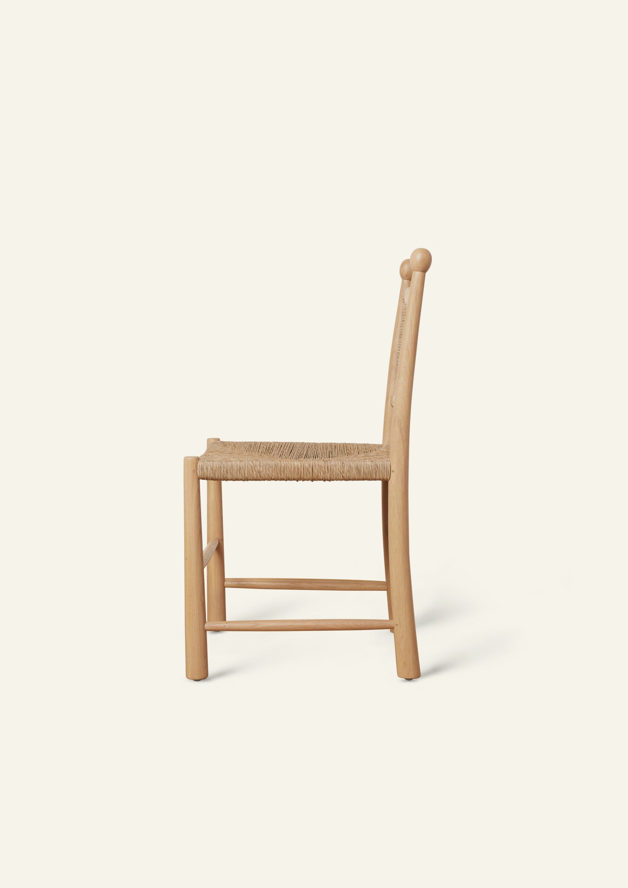 Side profile of a light oak dining chair with a handwoven rush seat and woven backrest. The chair features gently curved back posts, rounded legs, and simple stretcher supports, highlighting its handcrafted construction and clean silhouette against a soft ivory background.