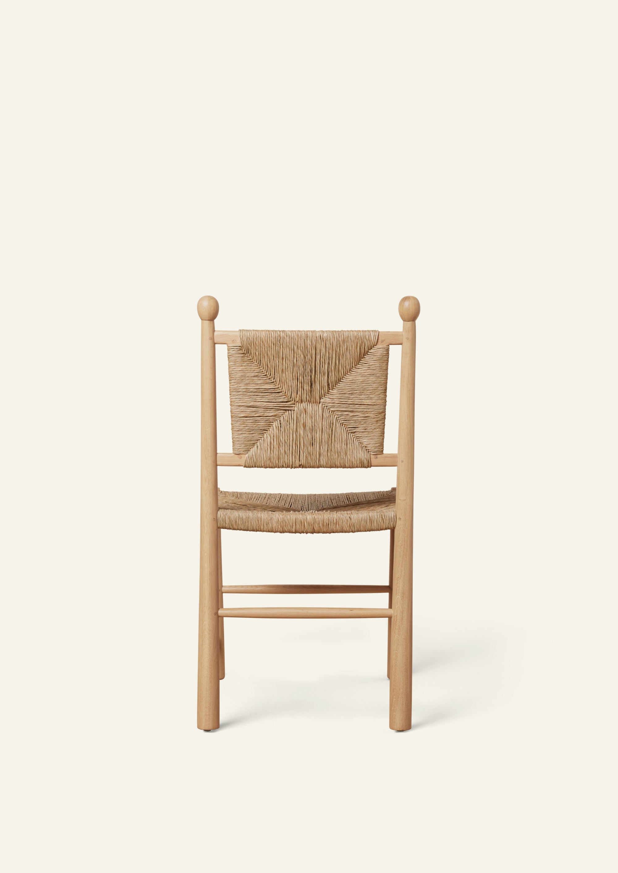 Rear view of a white oak dining chair with a handwoven rush seat and decorative woven backrest. Rounded back posts with turned finials frame the geometric rush pattern, while simple stretcher supports add stability. Photographed against a soft ivory background.