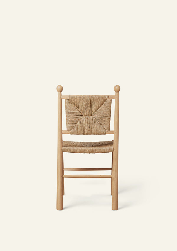 Rear view of a white oak dining chair with a handwoven rush seat and decorative woven backrest. Rounded back posts with turned finials frame the geometric rush pattern, while simple stretcher supports add stability. Photographed against a soft ivory background.