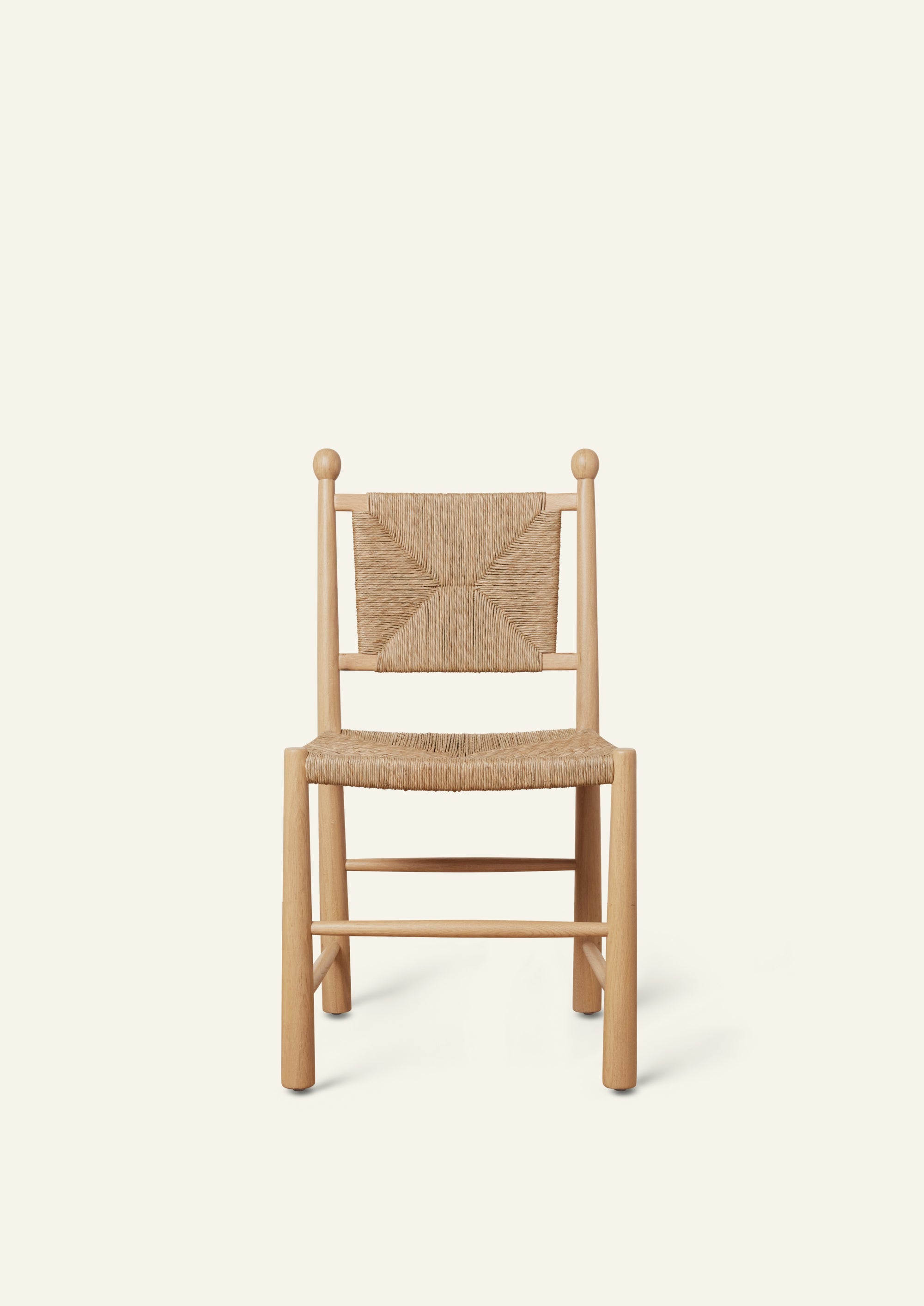 Front view of a white oak dining chair featuring a handwoven rush seat and geometric woven backrest. The chair has rounded legs, simple stretcher supports, and decorative finials atop the back posts, shown against a warm ivory backdrop.