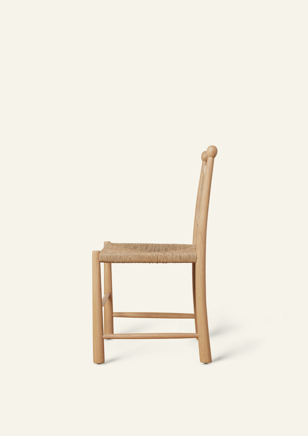 Side profile of a white oak dining chair with a handwoven rush seat and woven backrest. The chair features gently curved back posts, rounded legs, and simple stretcher supports, highlighting its handcrafted construction and clean silhouette against a soft ivory background.