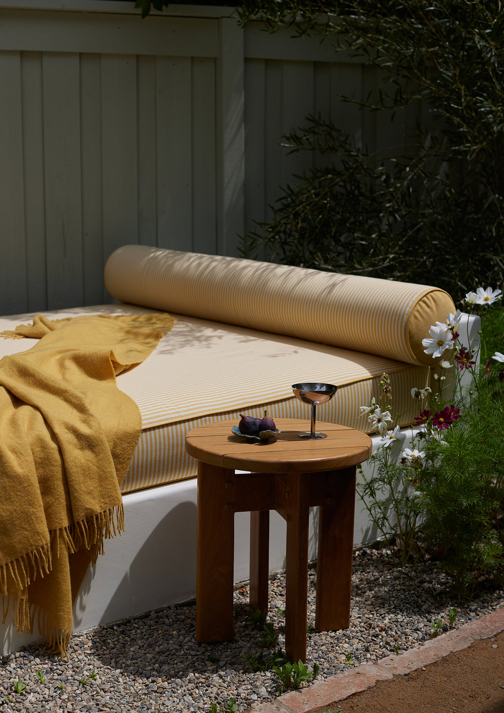 The Otto Side Table styled beside an outdoor daybed with striped cushions and a mustard throw. Its sculptural silhouette and solid teak construction add warmth and practicality to outdoor lounging spaces.