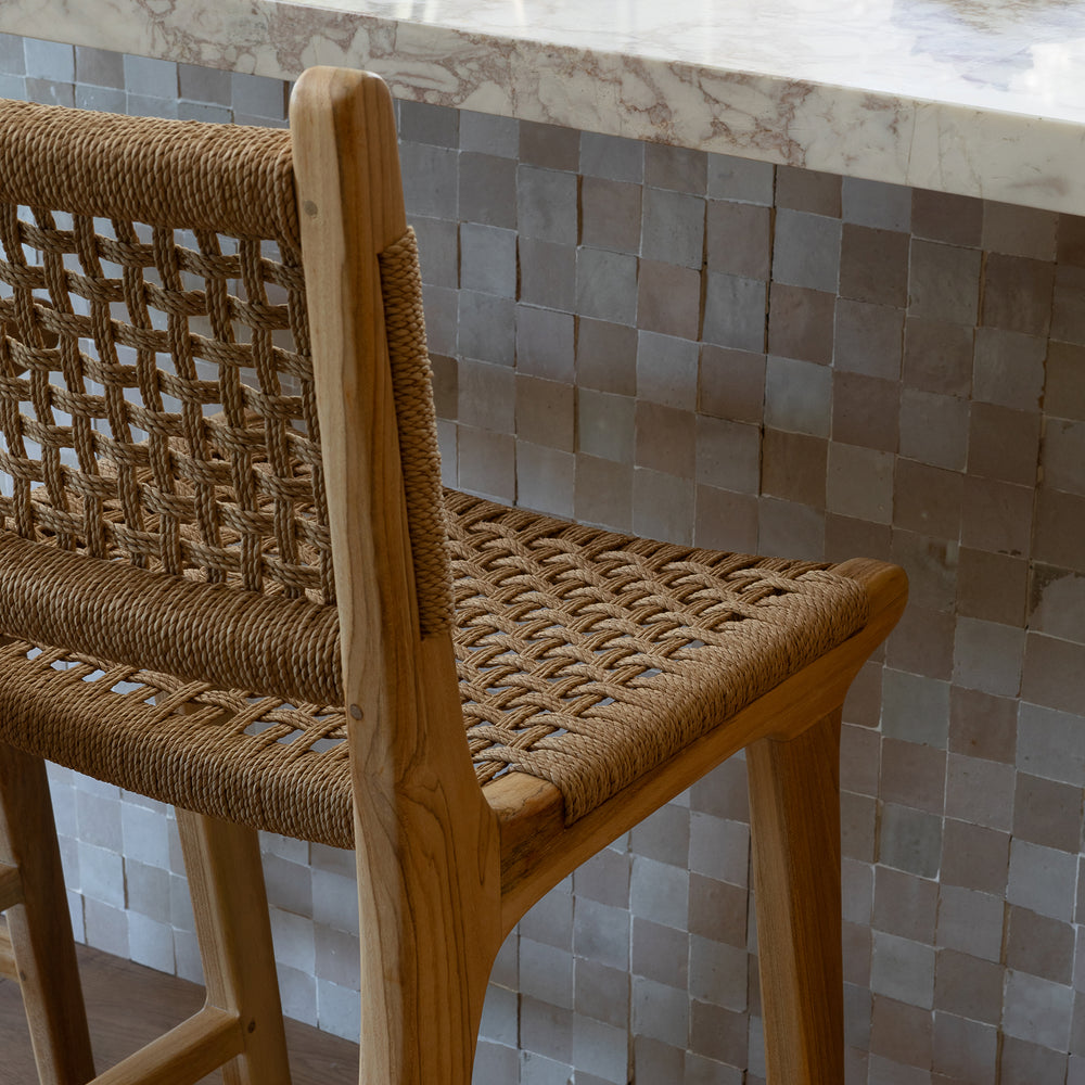 Wooden bar stool with woven seat and backrest against a tiled wall.