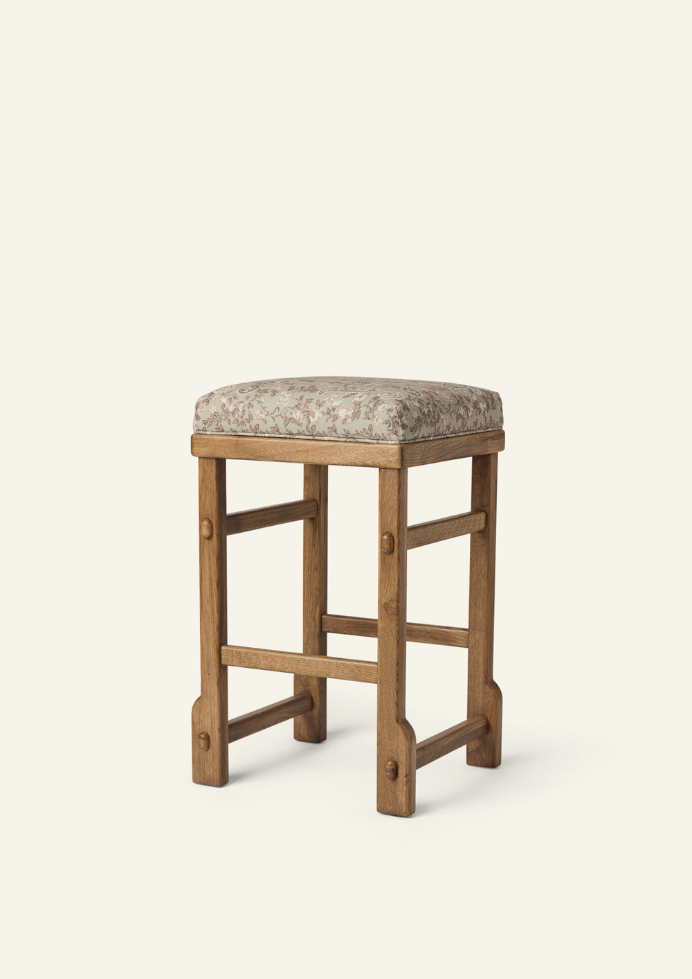 Upholstered counter stool featuring a softly cushioned seat in sage floral fabric, set atop a solid honey oak frame with exposed joinery details. The muted floral pattern adds a subtle, vintage-inspired touch to the timeless silhouette.
