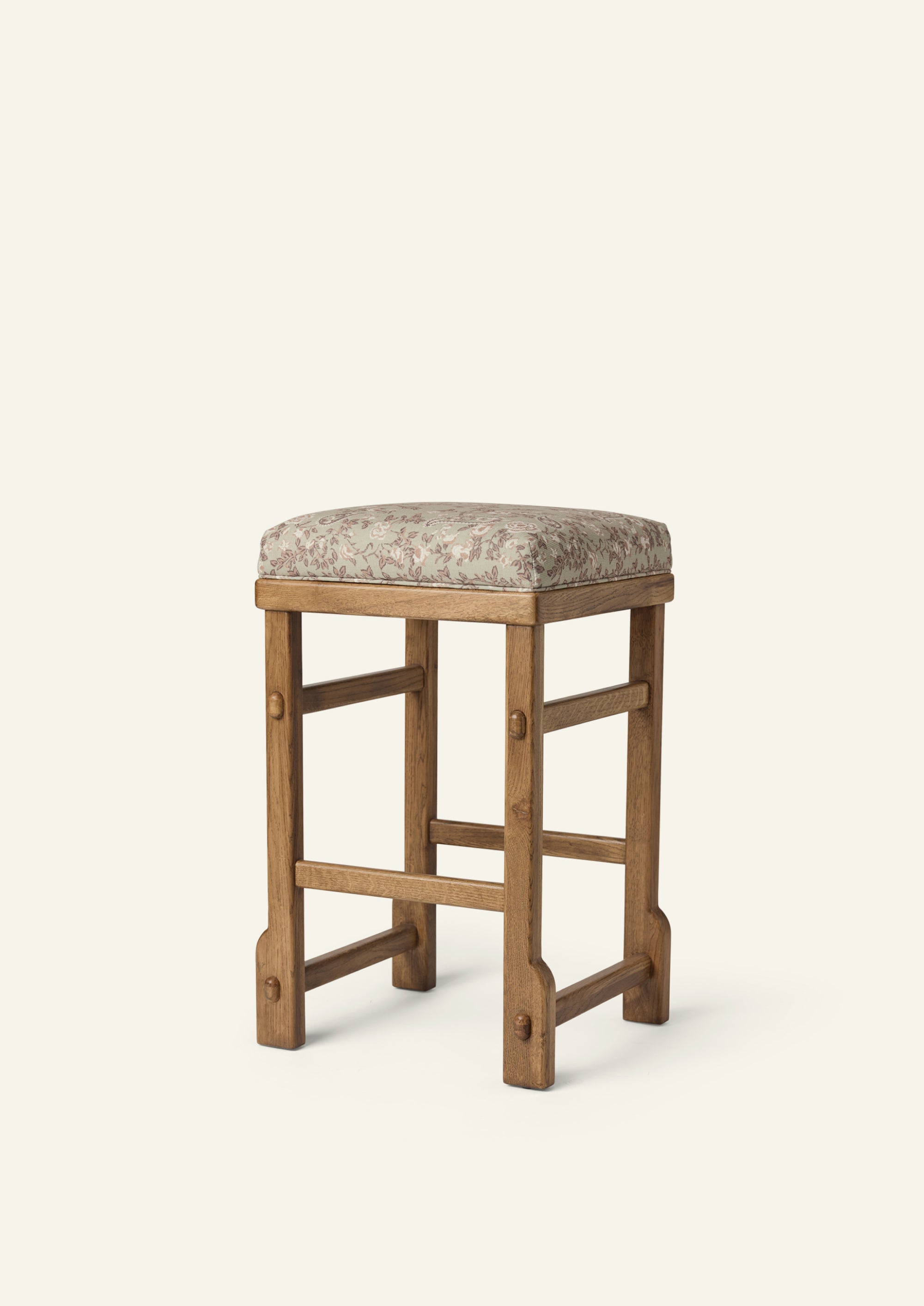 Upholstered counter stool featuring a softly cushioned seat in sage floral fabric, set atop a solid honey oak frame with exposed joinery details. The muted floral pattern adds a subtle, vintage-inspired touch to the timeless silhouette.