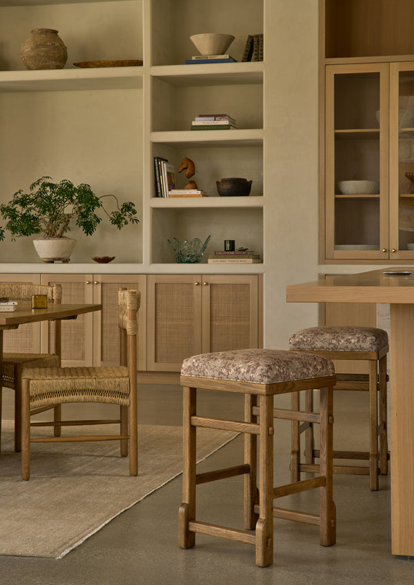 A pair of upholstered counter stools in Aloe Botanical Floral are styled alongside a light oak dining table in a warm, layered interior. The floral upholstery introduces softness and pattern while complementing the natural wood finishes throughout the space.