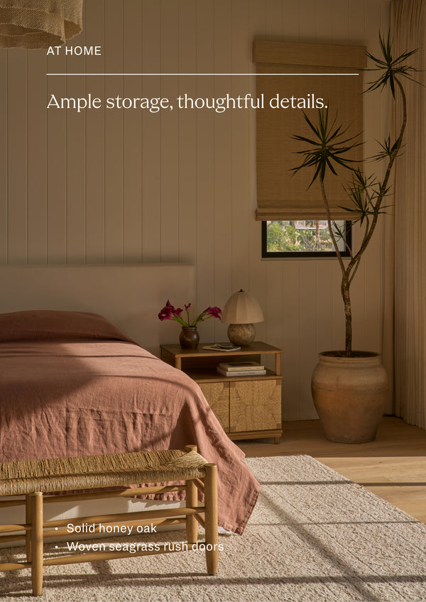 Ample storage, thoughtful details. Honey oak nightstand styled beside a bed in a warm, light-filled bedroom. The woven seagrass rush doors and open shelf create a functional storage solution with natural, organic appeal.