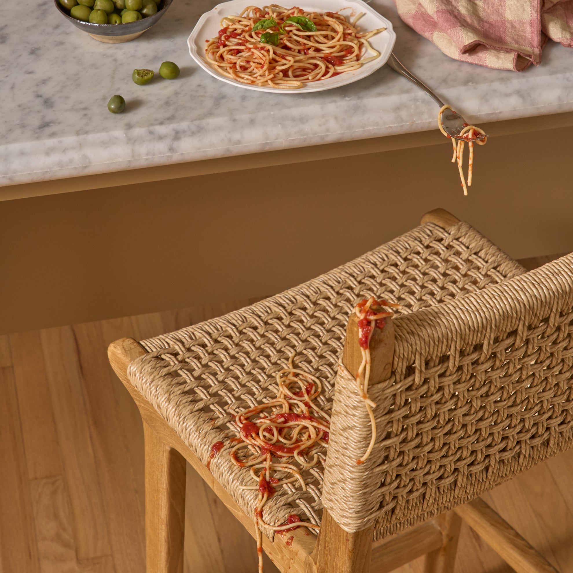 Synthetic Paper Cord Teak stool with spaghetti on a marble countertop with pasta and olives.