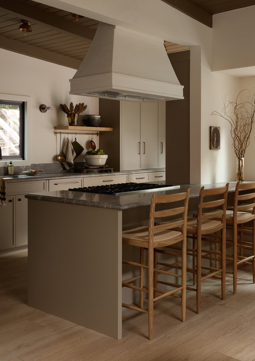 A modern greige European style kitchen with the Tiga honey oak counter stool with a paper cord seat. Soft lighting, creamy whites, and various kitchen decor make for a cozy feeling space. 