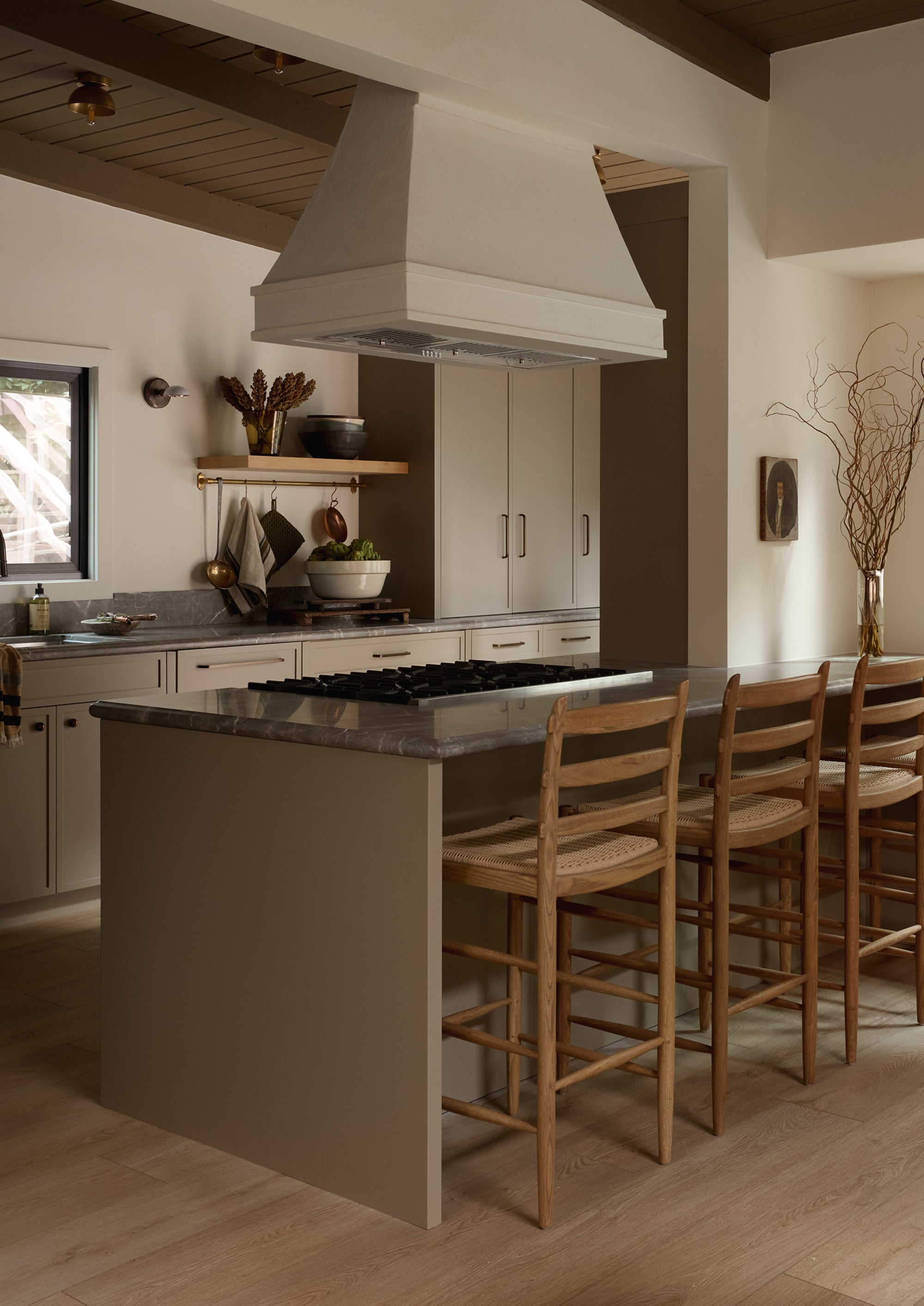 A modern greige European style kitchen with the Tiga honey oak counter stool with a paper cord seat. Soft lighting, creamy whites, and various kitchen decor make for a cozy feeling space. 