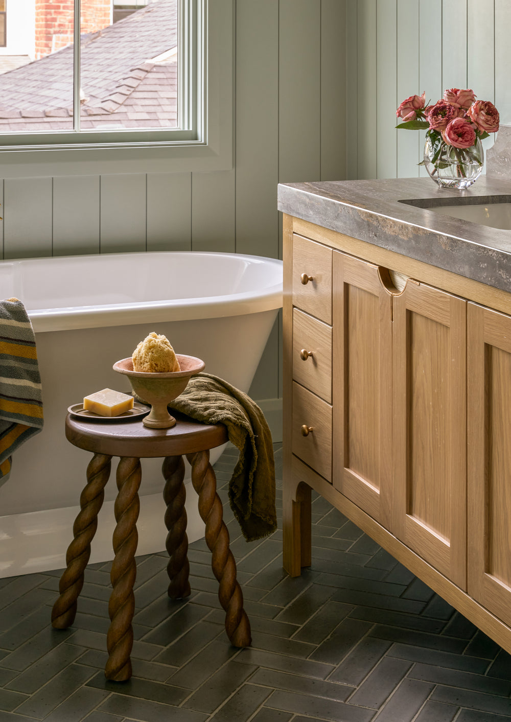 Round oak stool styled beside a freestanding bathtub and vanity, topped with bath essentials and a folded towel. The warm wood finish and twisted legs add charm and functionality to the bathroom setting.