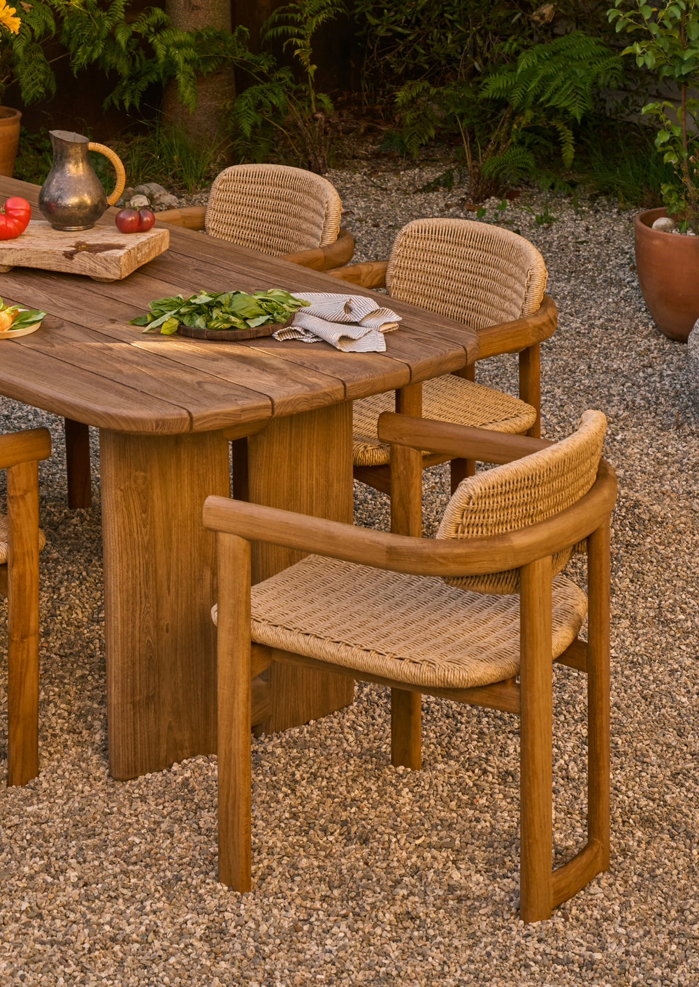 Teak dining armchairs arranged around an outdoor dining table in a garden setting. The woven textures and natural materials complement the surrounding landscape, creating an inviting space for gathering.