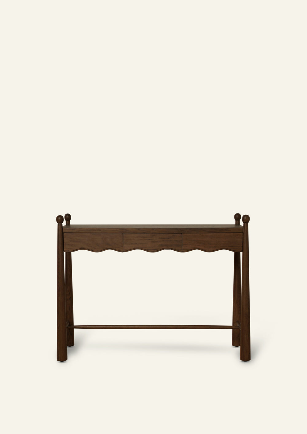 Front view of a dark oak console table featuring three drawers, a gently scalloped apron, and tapered legs topped with rounded finials. A lower stretcher shelf adds stability while highlighting the table’s traditional silhouette. Photographed against a soft ivory background.
