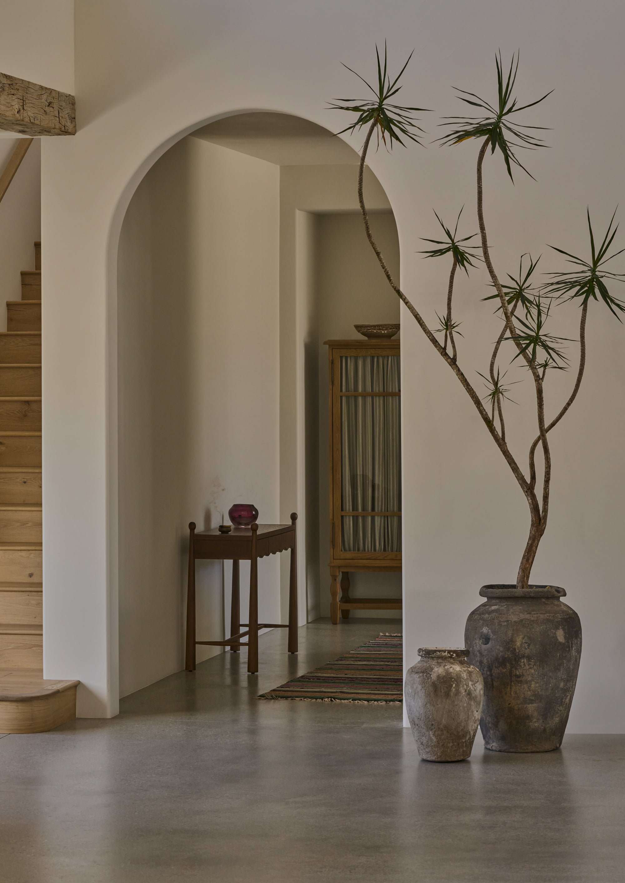 Dark oak console table styled in a hallway beneath a rounded archway. Decorative objects rest on the tabletop while oversized ceramic vessels and a sculptural indoor tree create a warm, collected entryway setting.