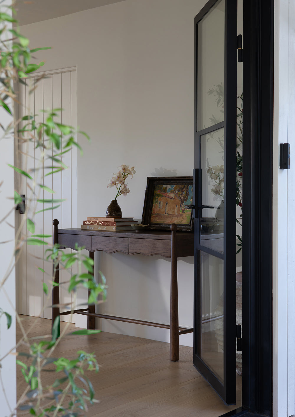 Dark oak console table styled in an entryway and viewed through an open glass door. Stacked books, framed artwork, and fresh flowers create a welcoming, lived-in vignette.