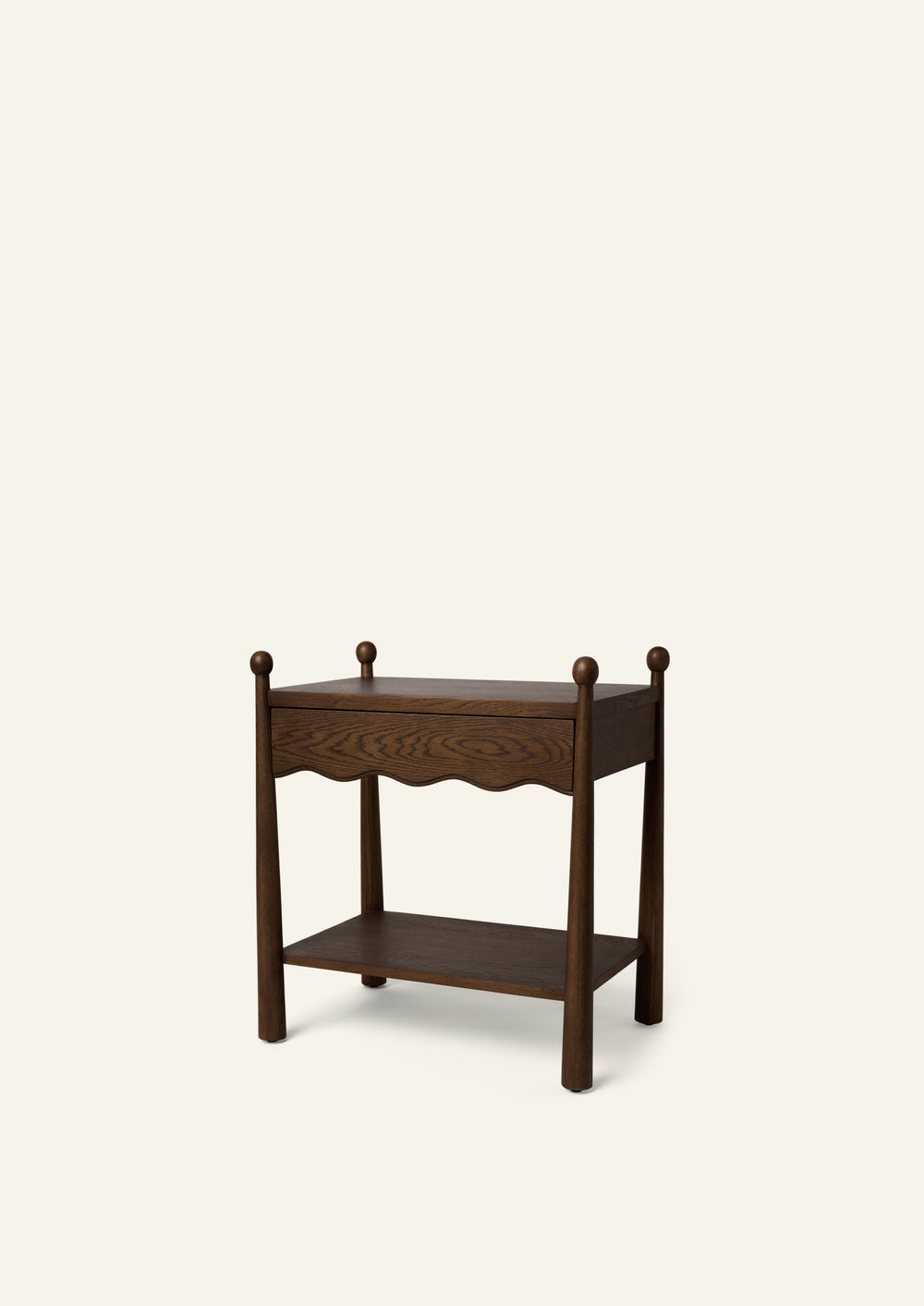 Three-quarter view of a dark oak nightstand featuring a single drawer, open lower shelf, rounded finials, and a softly scalloped apron. The rich wood grain and traditional detailing are highlighted against a warm ivory background.