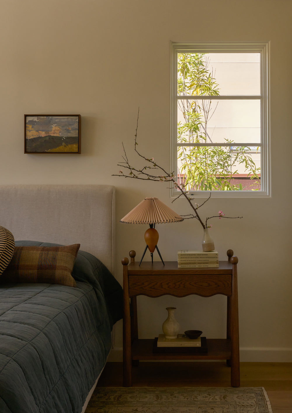 Dark oak nightstand styled beside an upholstered bed beneath a sunlit window. Decorative books, a pleated table lamp, and flowering branches create a calm, layered bedroom setting.