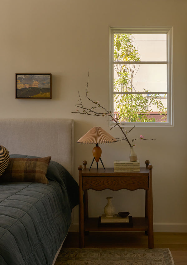 Dark oak nightstand styled beside an upholstered bed beneath a sunlit window. Decorative books, a pleated table lamp, and flowering branches create a calm, layered bedroom setting.