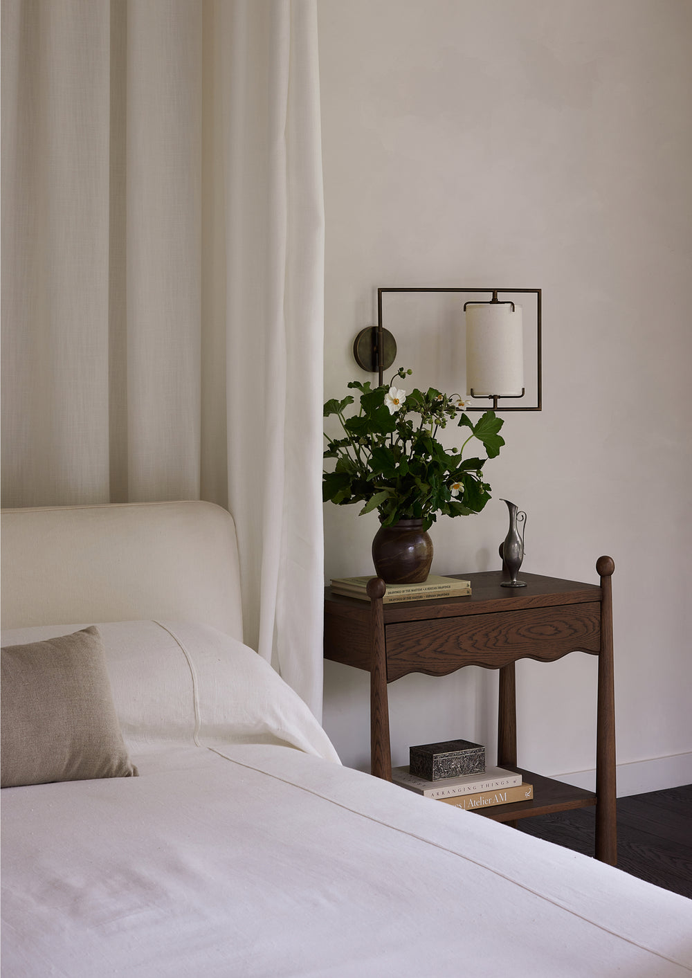 Dark oak nightstand styled beside a bed with crisp white linens. A vase of fresh flowers, books, and decorative objects rest on the tabletop, creating a warm and inviting bedroom vignette.