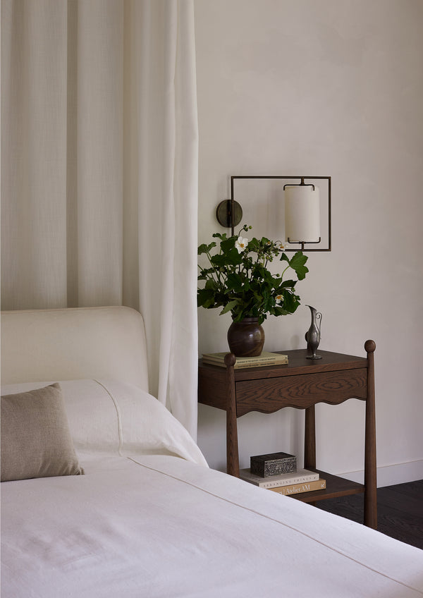 Dark oak nightstand styled beside a bed with crisp white linens. A vase of fresh flowers, books, and decorative objects rest on the tabletop, creating a warm and inviting bedroom vignette.