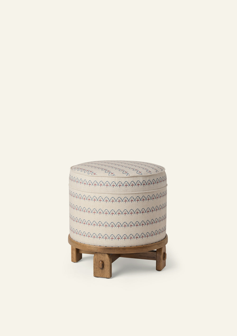 Round upholstered pouf in the Neutral Arches fabric, featuring a cream background with blue and dusty rose scalloped motifs. The pouf sits on a sculptural honey oak base with exposed joinery details.
