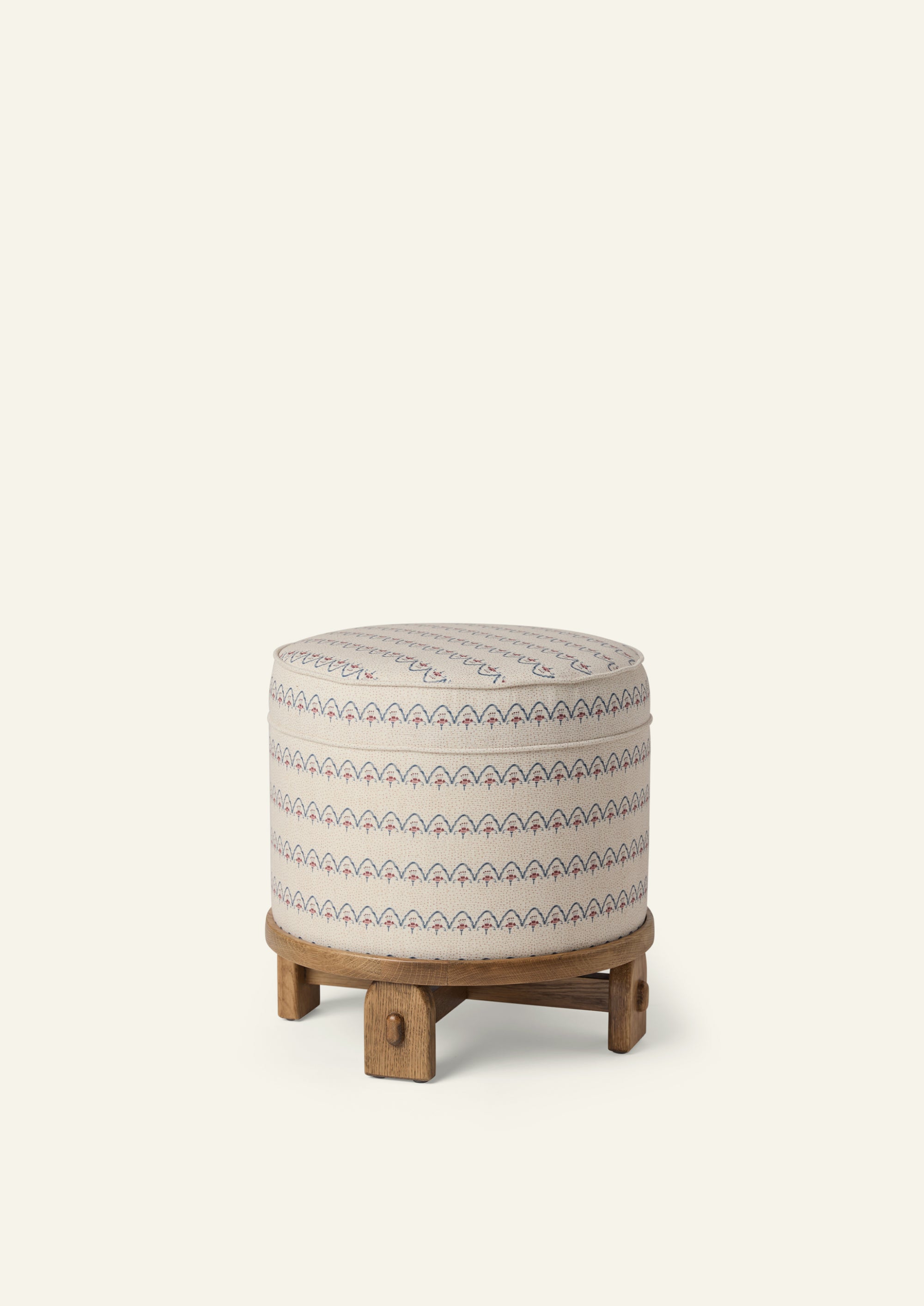 Round upholstered pouf in the Neutral Arches fabric, featuring a cream background with blue and dusty rose scalloped motifs. The pouf sits on a sculptural honey oak base with exposed joinery details.