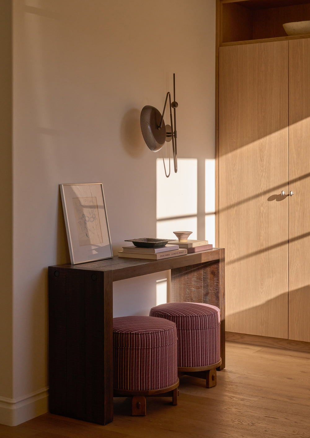 Two Yara Poufs in Oxblood Stripe are tucked beneath a dark wood console table in a warmly lit hallway. The compact upholstered stools provide versatile extra seating while adding color, pattern, and texture to the space.