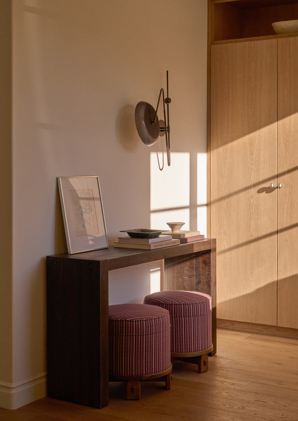 Two Yara Poufs in Oxblood Stripe are tucked beneath a dark wood console table in a warmly lit hallway. The compact upholstered stools provide versatile extra seating while adding color, pattern, and texture to the space.
