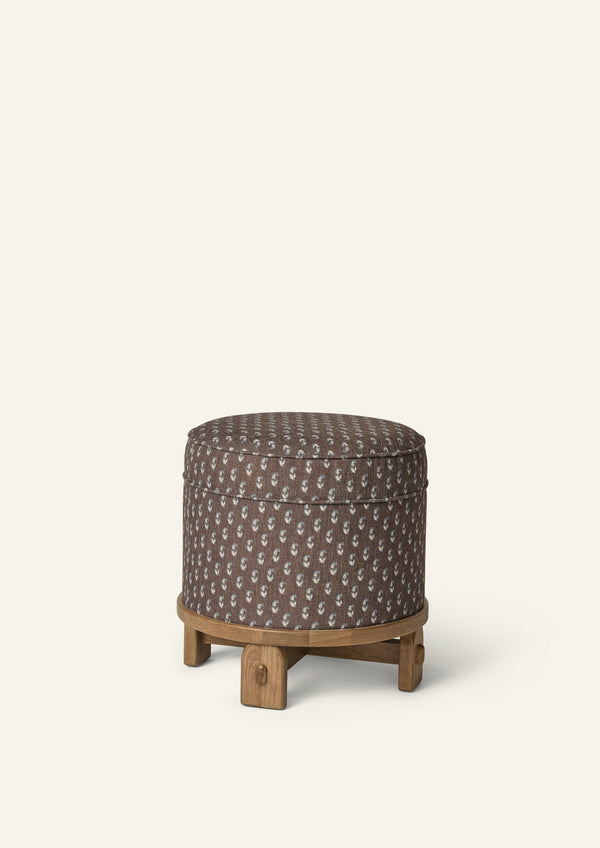 Round upholstered pouf in Cocoa Floral fabric, featuring a rich brown ground with a delicate scattered floral motif in soft blue and cream. The pouf sits atop a sculptural honey oak base with exposed joinery details.
