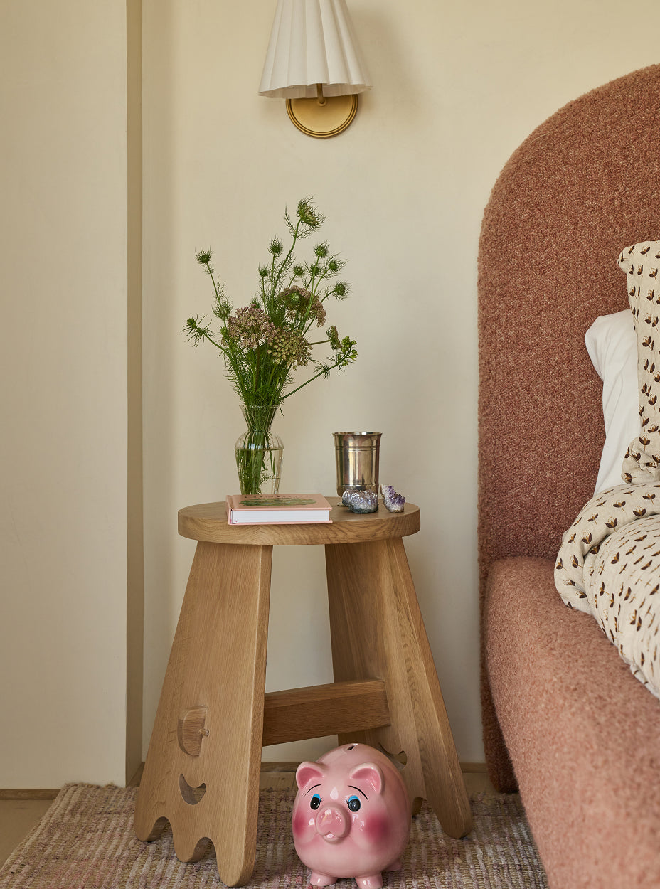 The senja side table with a vase of flowers, a book, and a piggy bank next to a textured headboard.