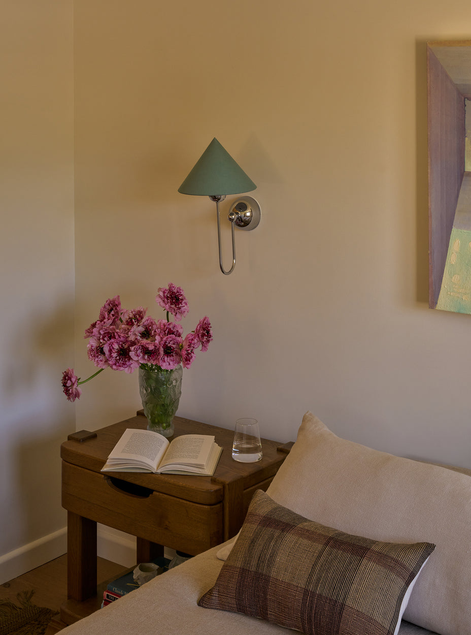 The mina nightstand with flowers and book next to a sofa in a room with a wall lamp.