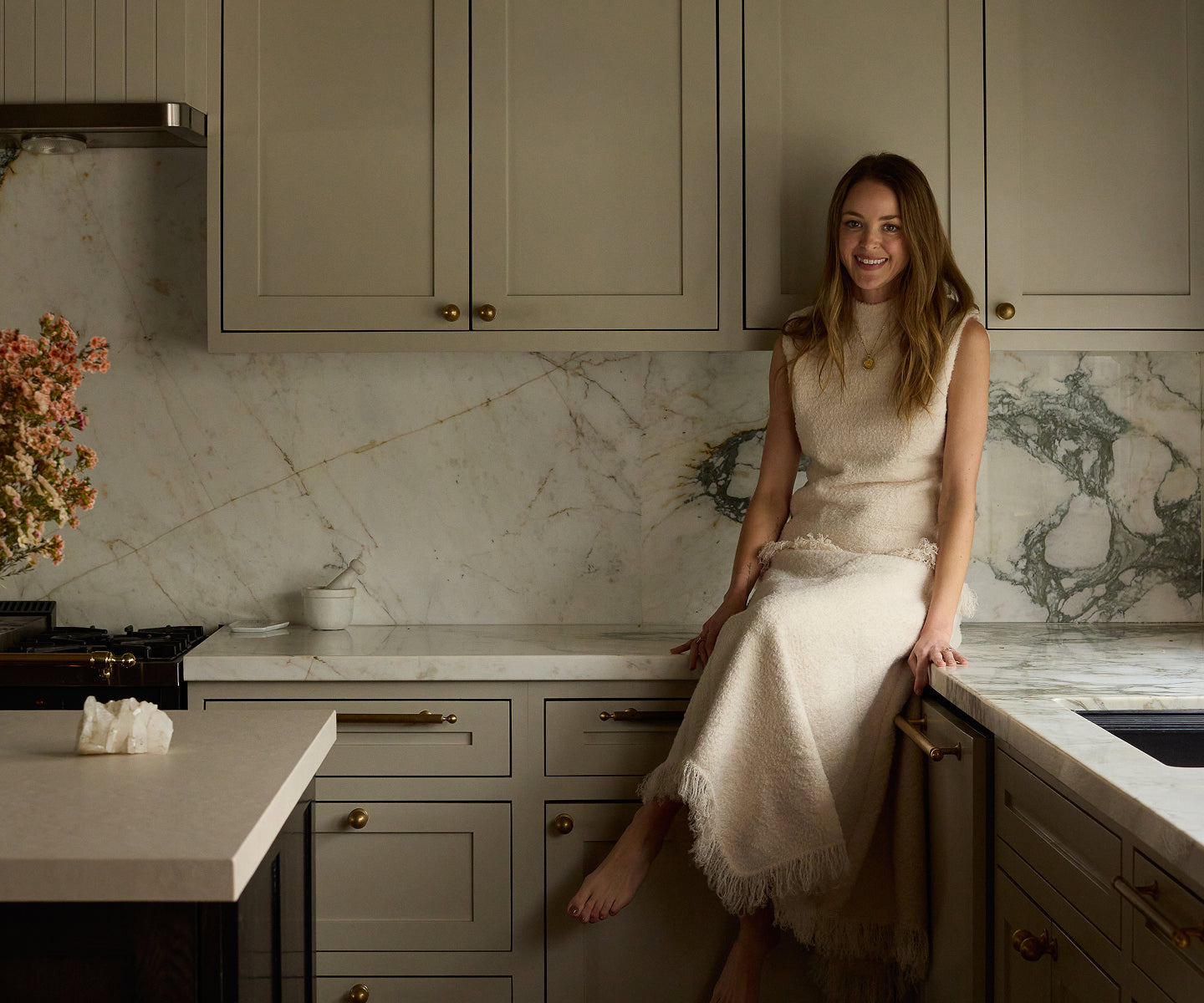 Kaitlynn Carter in a white dress sitting on a kitchen counter with marble backsplash