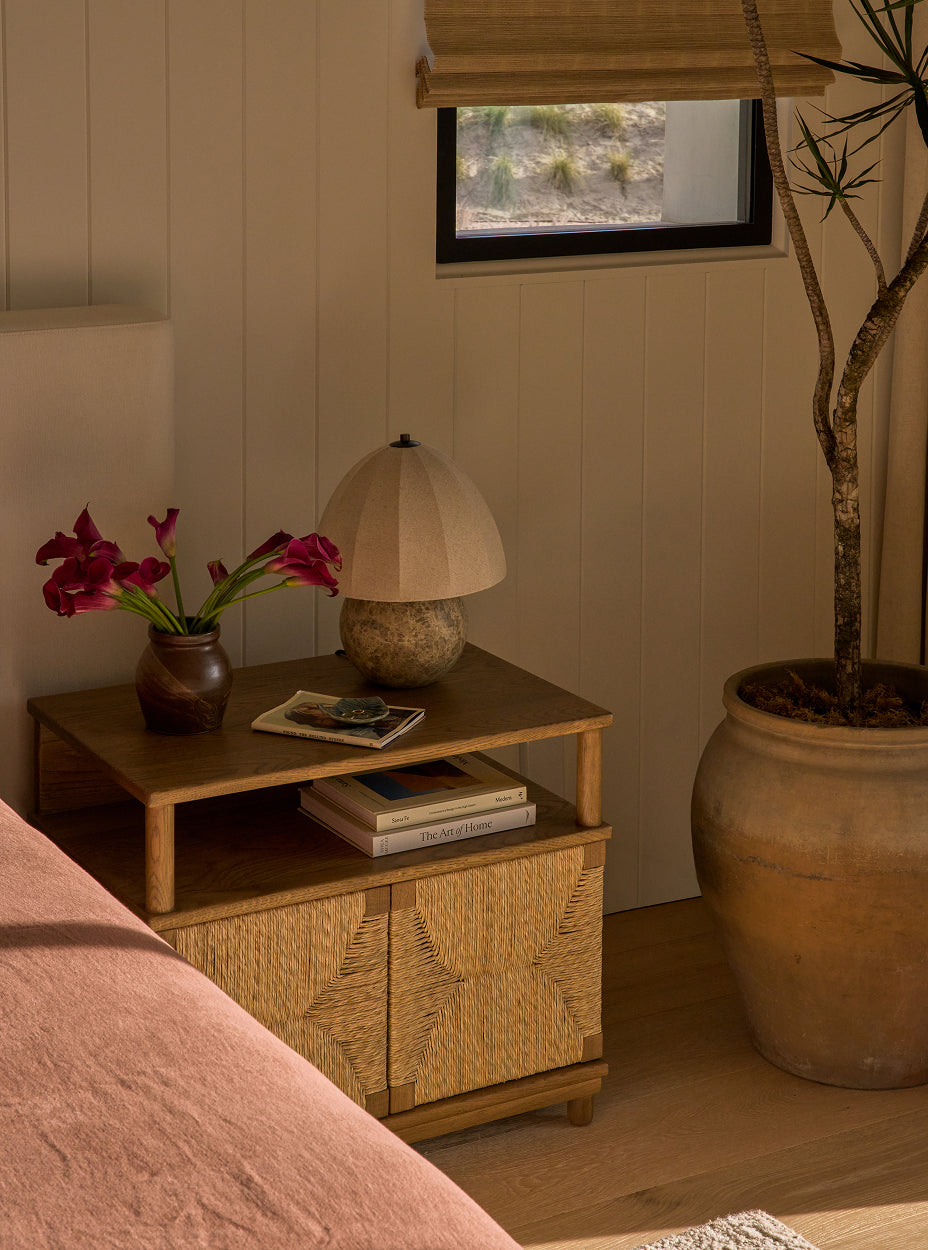 The suma nightstand with lamp, books, and flowers next to a large potted plant in a room.