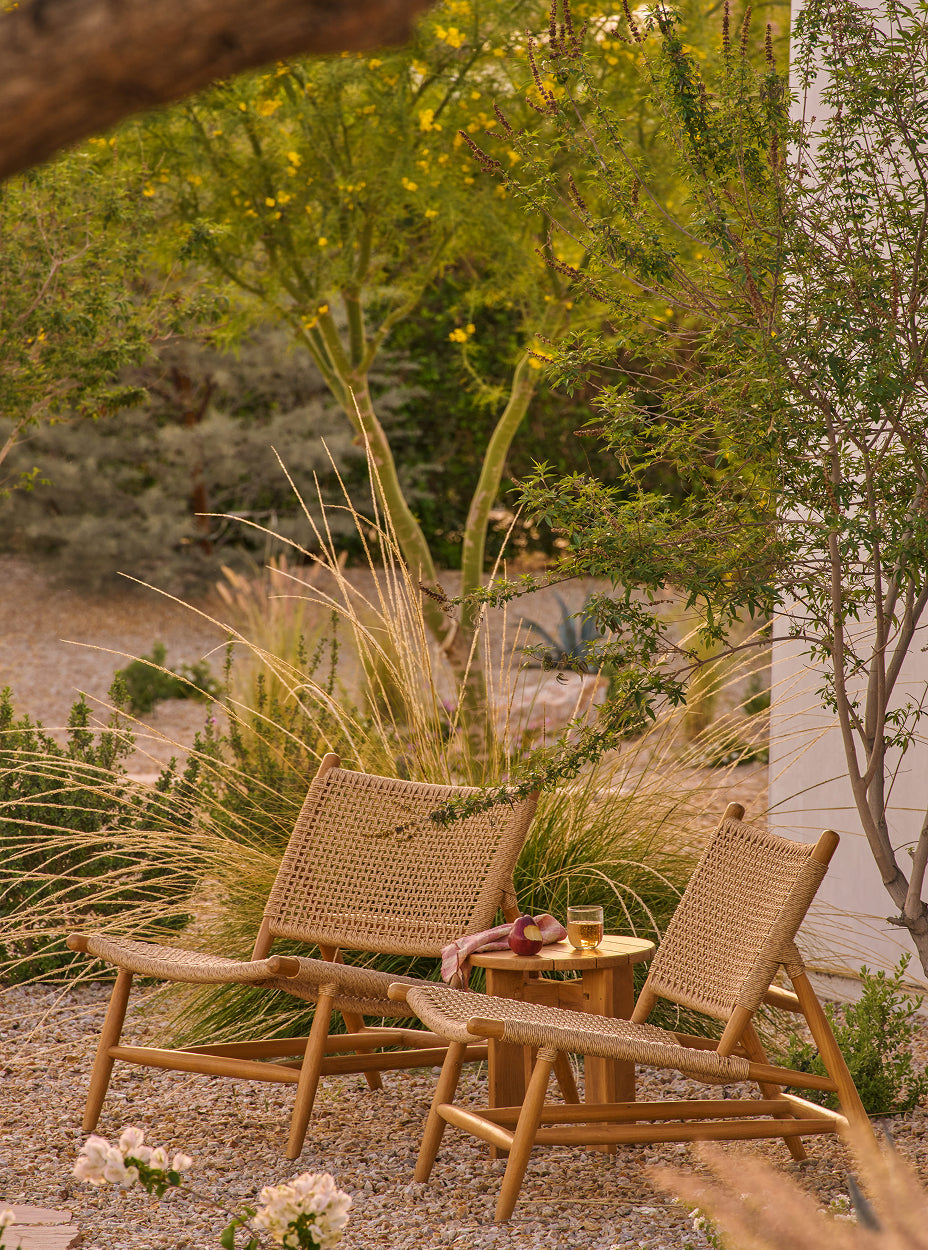 Two bali lounge chairs with a small table in a natural setting with trees and plants.