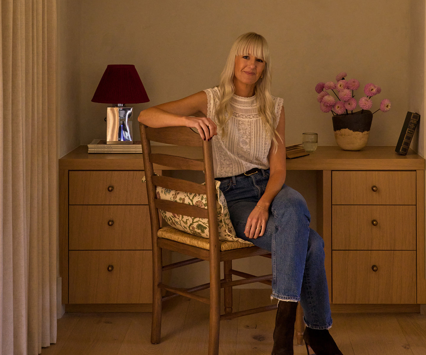 Kelli Murray sitting on a wooden chair in a room with a lamp and a vase of flowers.