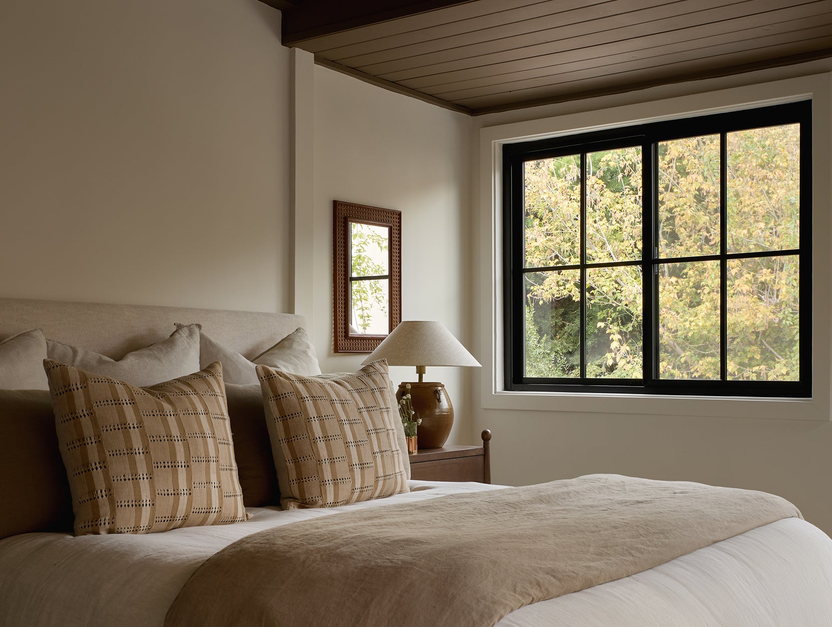 Bedroom with a bed featuring plaid pillows, a lamp on Wave nightstand, and a window with a view of trees.