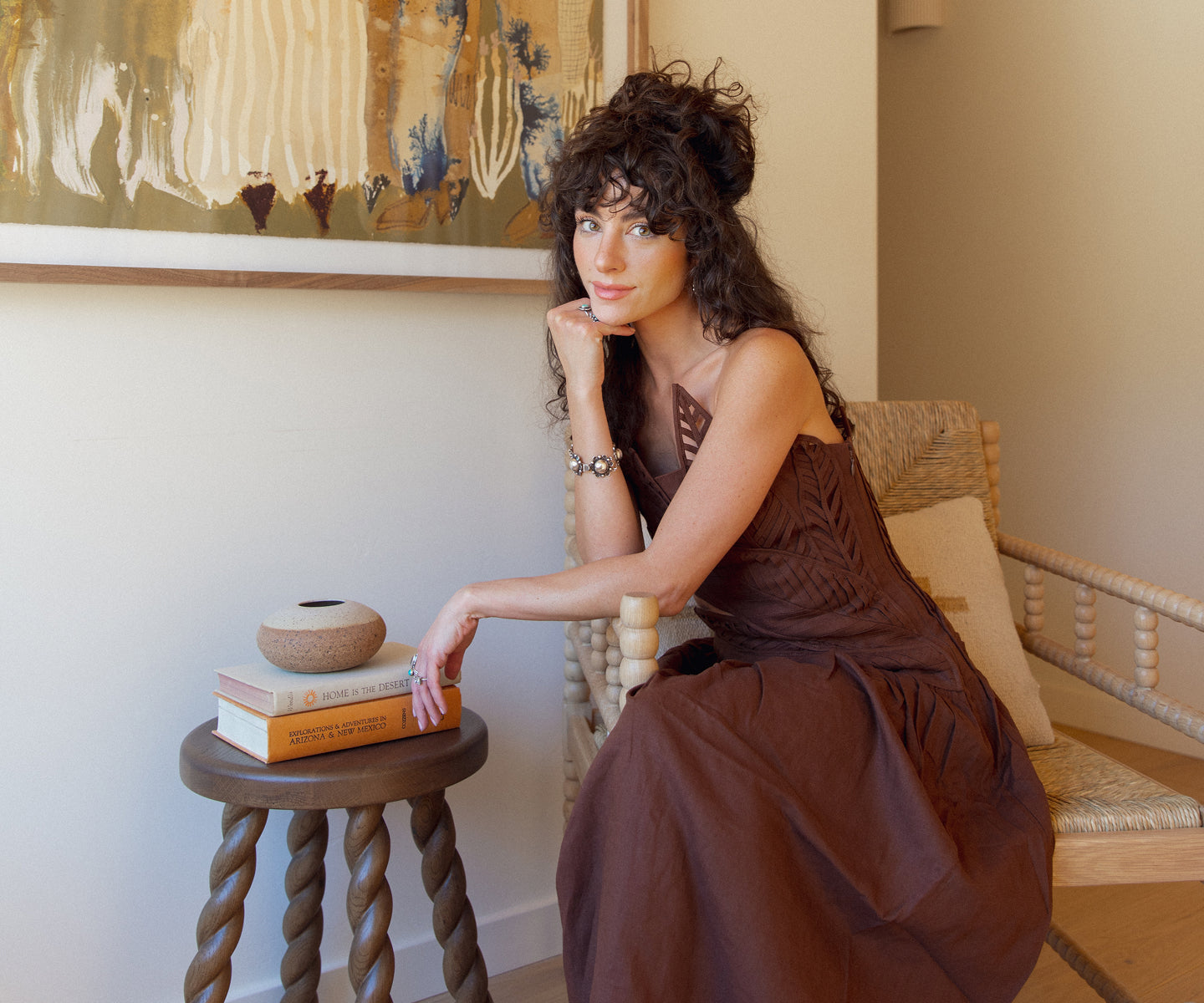 New Darlings in a brown dress sitting on a chair in a room with a painting and books.