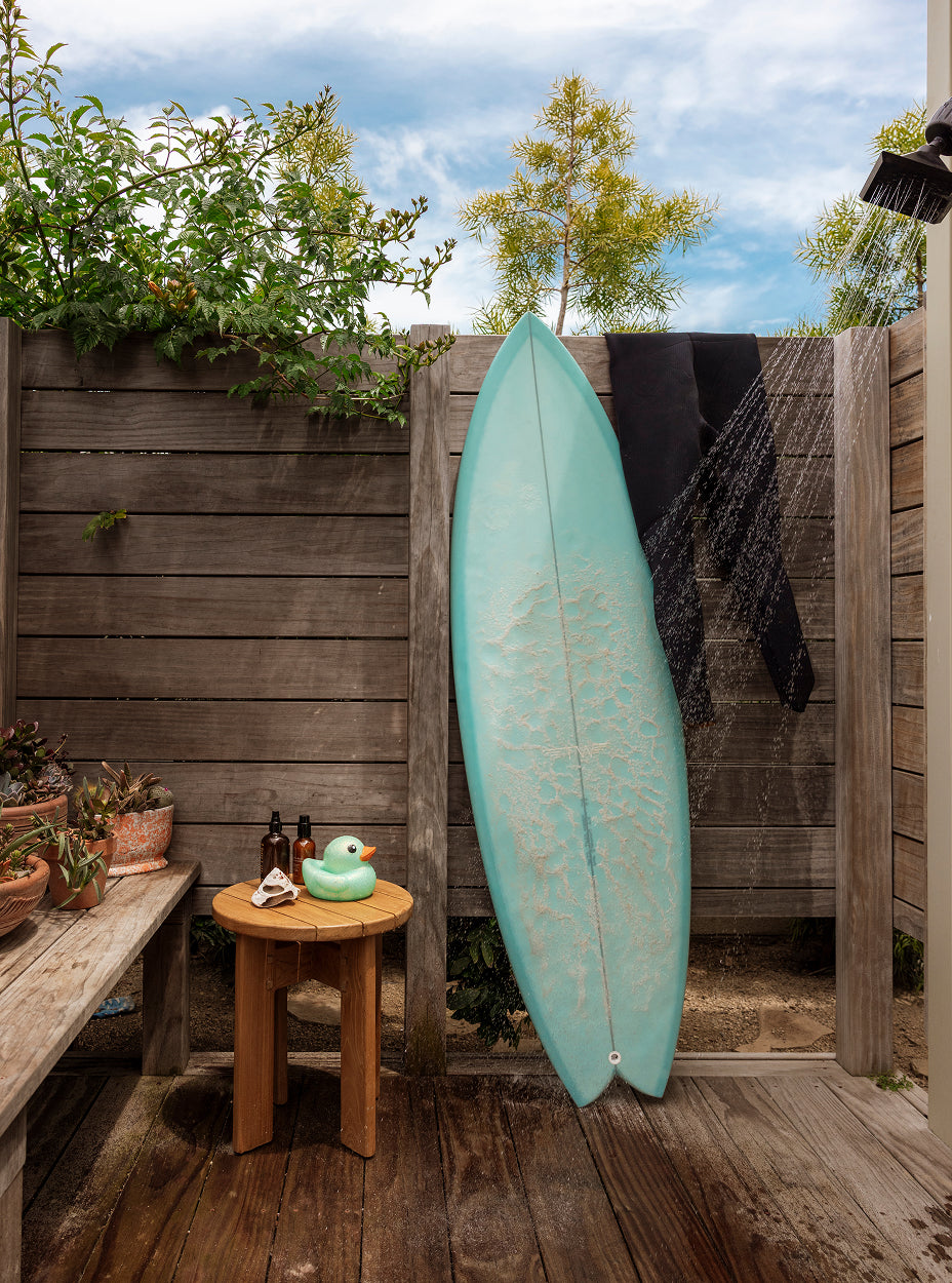 Teal surfboard leaning against a wooden fence with a otto side table and plants in the foreground.