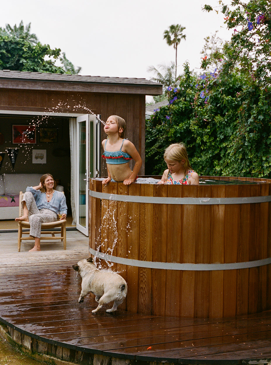 Children playing in a wooden hot tub with a dog, surrounded by greenery and a house in the background.